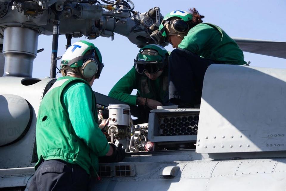 A group of three people dressed in green long sleeves look into the machinery of a helicopter rotor.