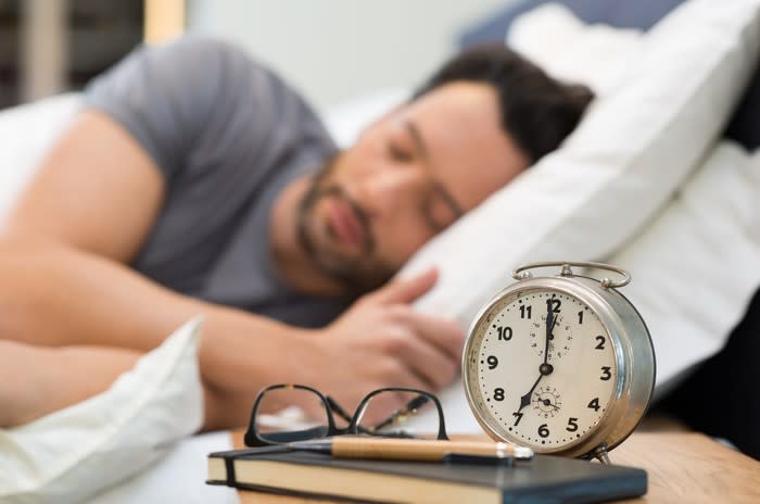 A man sleeping with an alarm clock and glasses in the foreground.
