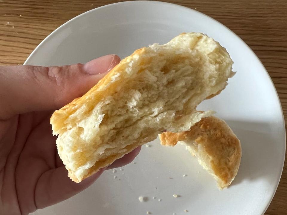 A hand holds a biscuit split in half above a white plate on a wood surface.