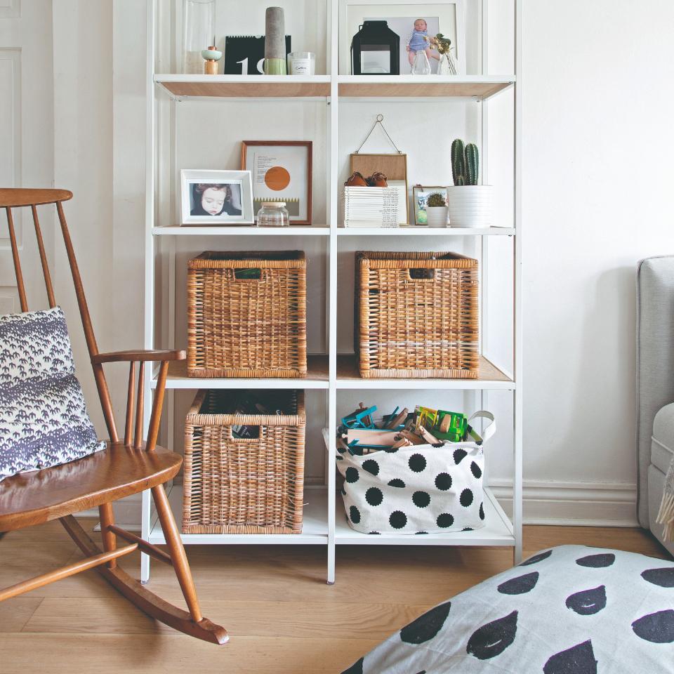 A living room with a white metal bookshelf displaying photos and decor, along with wicker storage baskets with a wooden rocking chair next to it