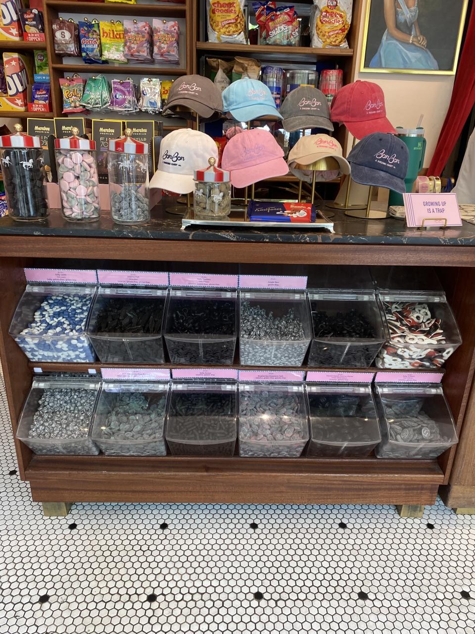 A candy shop display features various clear bins filled with different types of candies, jars of lollipops, and a selection of branded baseball caps on top