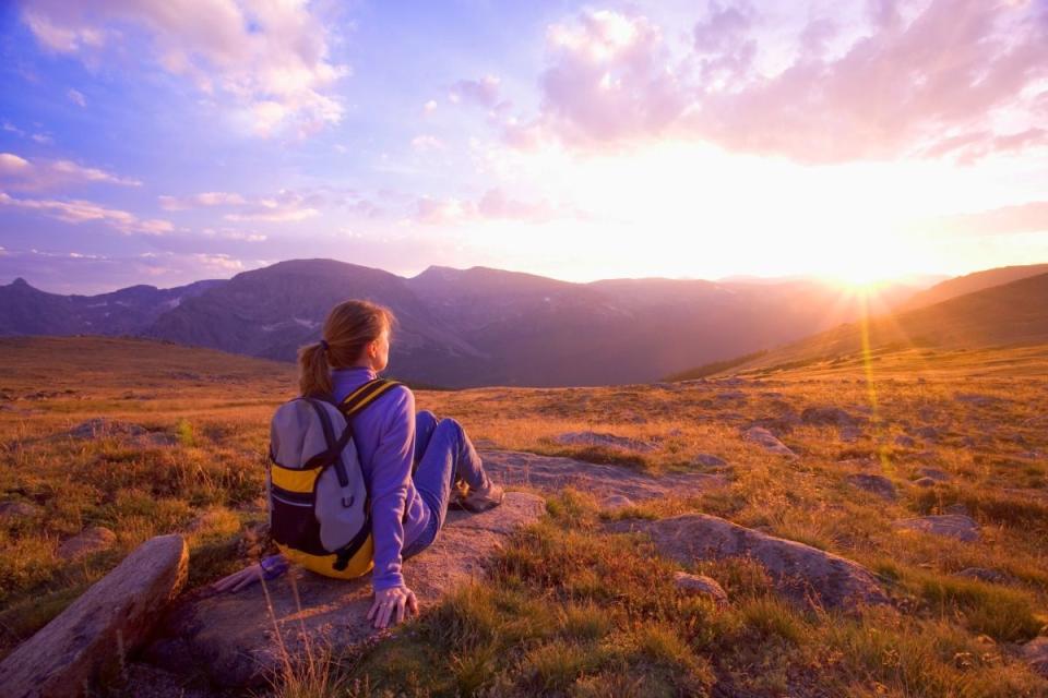 a women wearing a backpack sits on a rock watching a sunset over the mountains, a summer bucket list idea