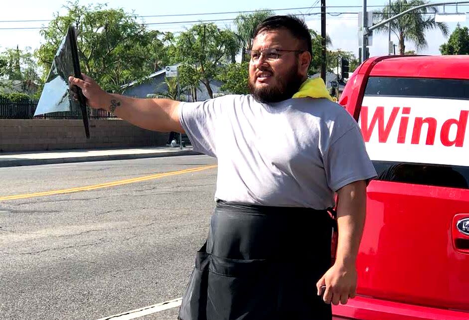 A man in a work apron standing by a parked car, waving a piece of window tint