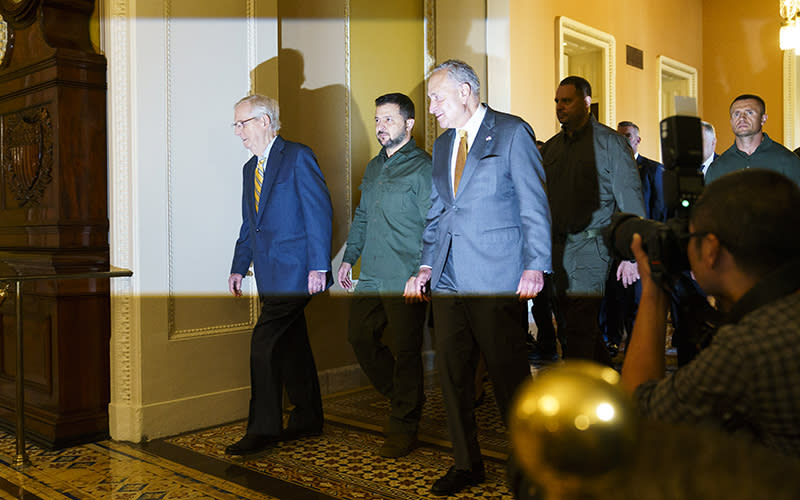 Ukrainian President Volodymyr Zelensky leaves an all-Senators meeting with Minority Leader Mitch McConnell (R-Ky.) and Majority Leader Chuck Schumer (D-N.Y.). The three men walk in front of a small group of men, some of whom are wearing suits while others wear green fatigues. A bright light shines toward the leaders, illuminating them in the dim hallway. To the right is a photographer, crouching to take a photo.