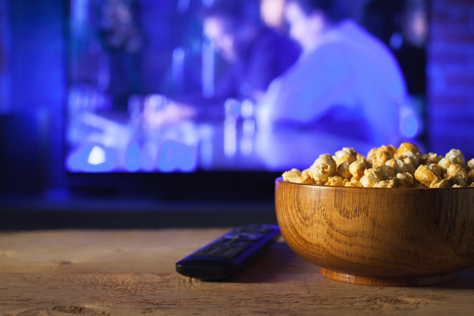 A wooden bowl of popcorn and the TV remote in the background the TV works. Evening cozy watching a movie or TV series at home.