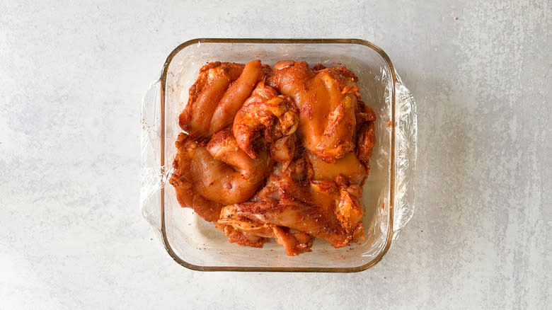 Chicken thighs with dry rub in glass baking dish covered with plastic wrap