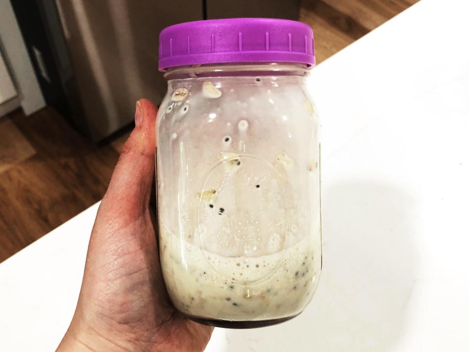 A hand holding a mason jar with a purple lid. Inside the jar is a mixture of shaken overnight oats.