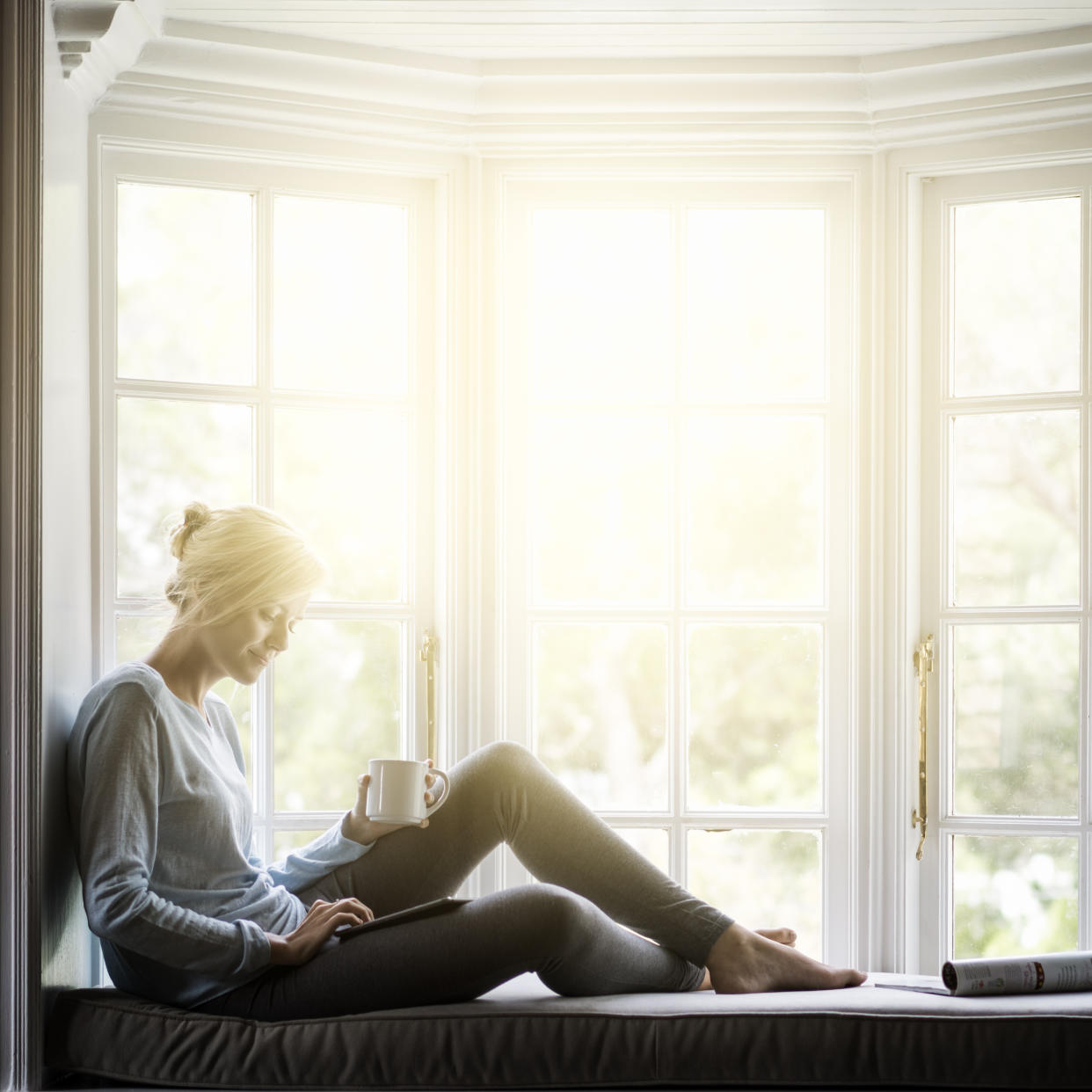 Junge Frau sitzt bei Sonne am Fenster und liest in einem Tablet