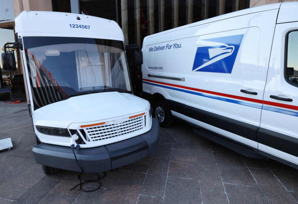 Two new USPS electric delivery vehicles parked side by side. One vehicle has "We Deliver For You" and the USPS logo on its side