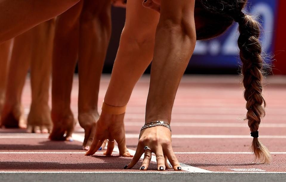 A closeup of women's hands, fingertips pressed into the ground, and one woman's braid, at the starting line.