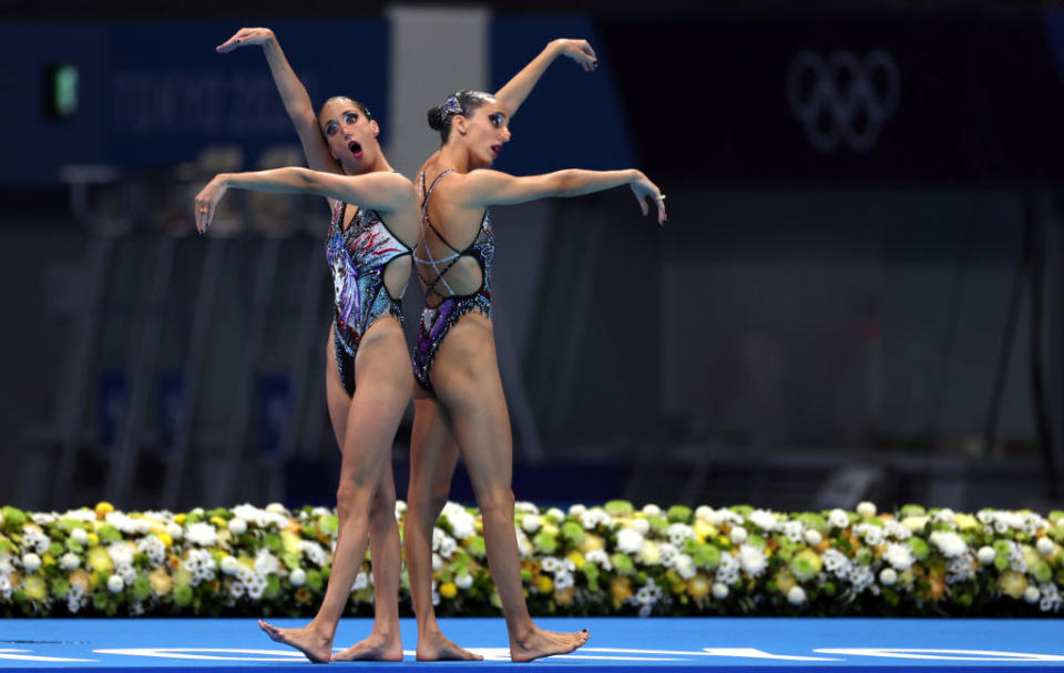 Two swimmers standing with their backs touching, arms raised the same way but seemingly haphazardly, and their mouths open