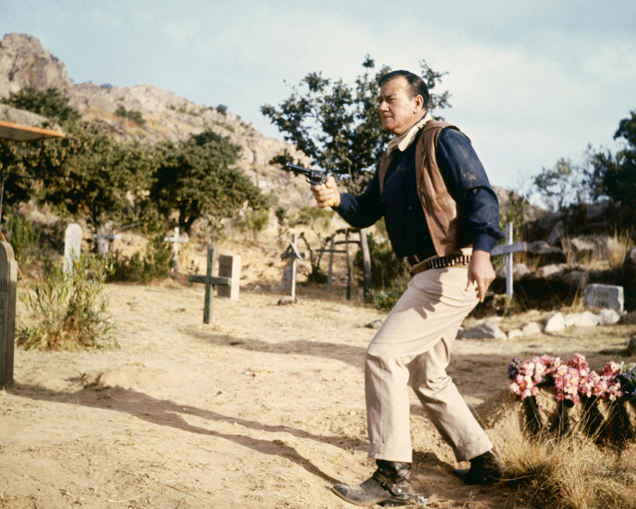 John Wayne (1907 - 1979), US actor wearing a tan leather waistcoat, a dark blue shirt and beige trousers, firing a pistol in a cemetery, circa 1960. (Photo by Silver Screen Collection/Getty Images)