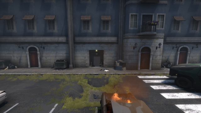 Video game screenshot from &#x002018;Counter-Strike: Global Offensive.&#x002019; On the side of a war-torn building is a dark passageway with a light over its entrance. A flaming car sits in the foreground.