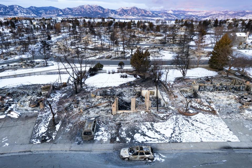 A burned neighborhood in ruins with mountains in the background. House foundations, a few brick walls and and shells of burned-out vehicles remain.