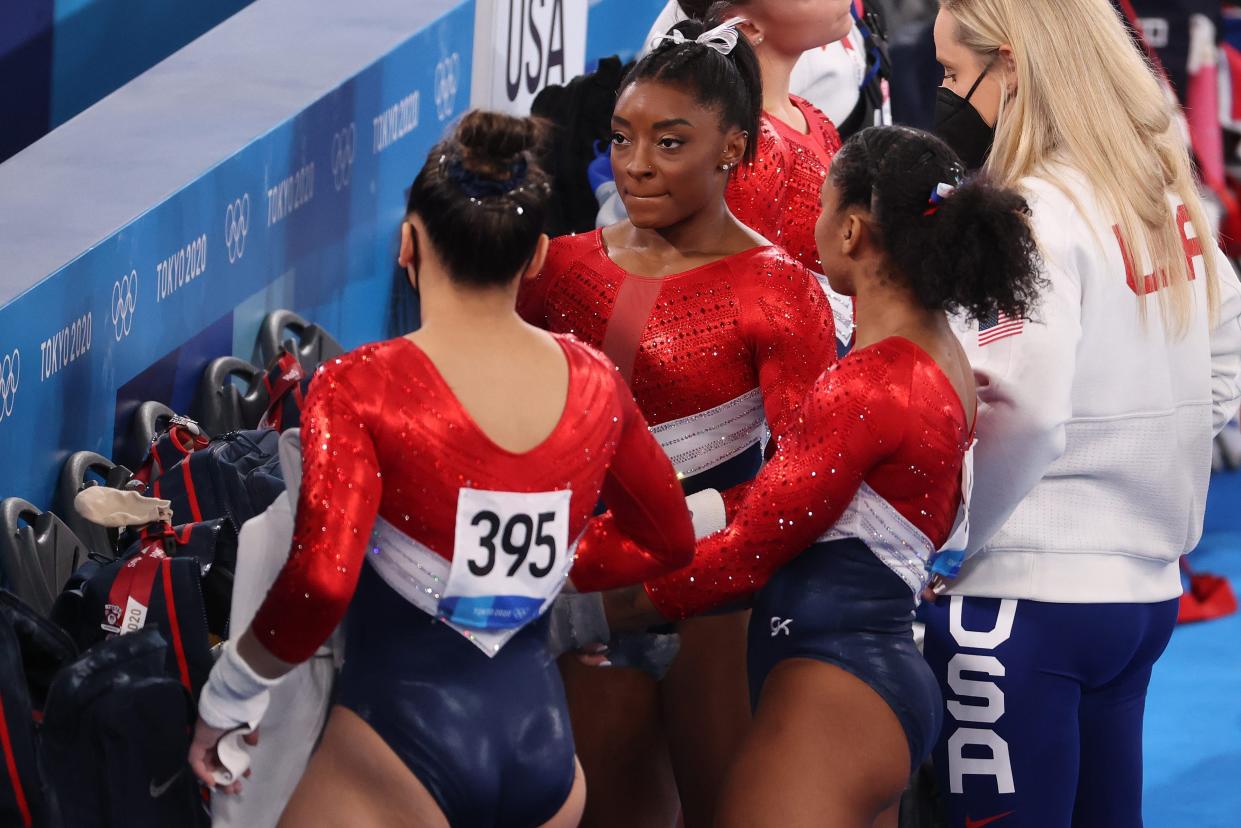 Biles, standing with two of her gymnastics teammates, is seen on the sidelines awaiting her score.