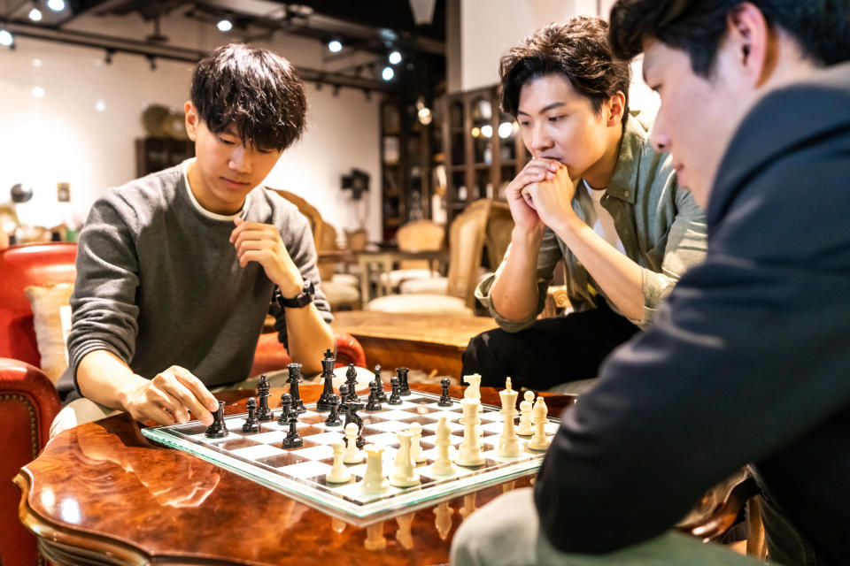 Three men concentrate on an exciting game of chess in a cozy, well-lit room with wooden furniture. Their expressions reflect deep concentration