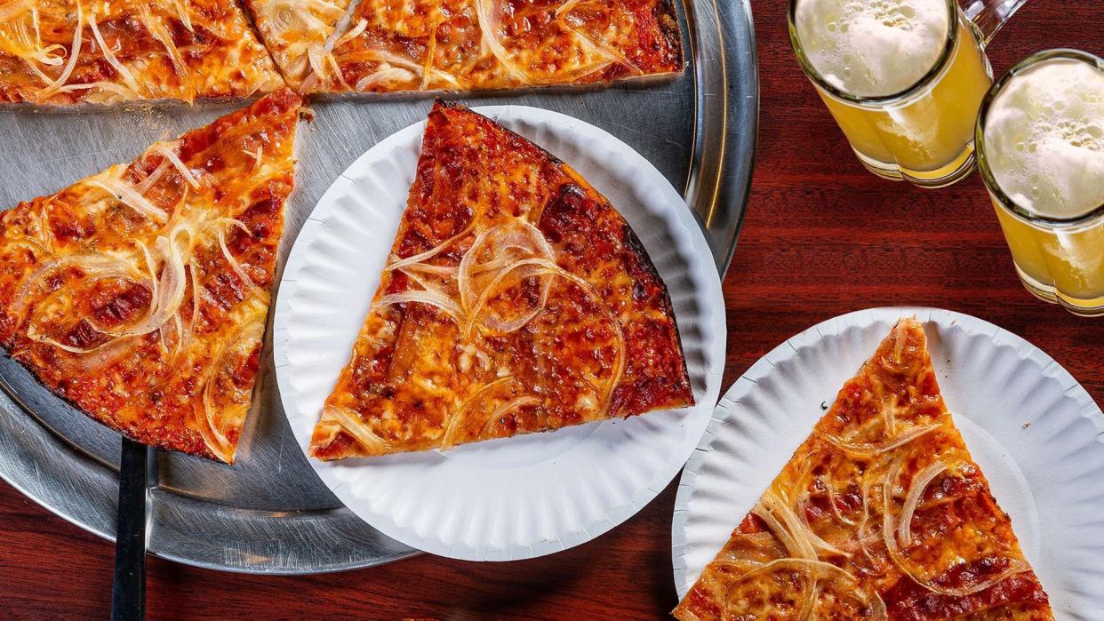 Top-view of slices of onion pizza, Colony Grill, Stamford, Connecticut, being cut from pizza dish and two paper plates, on a wooden table with two mugs of beer