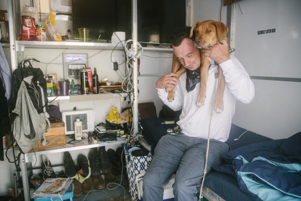 A puppy is draped around a man's neck and they are surrounded by his belongings.