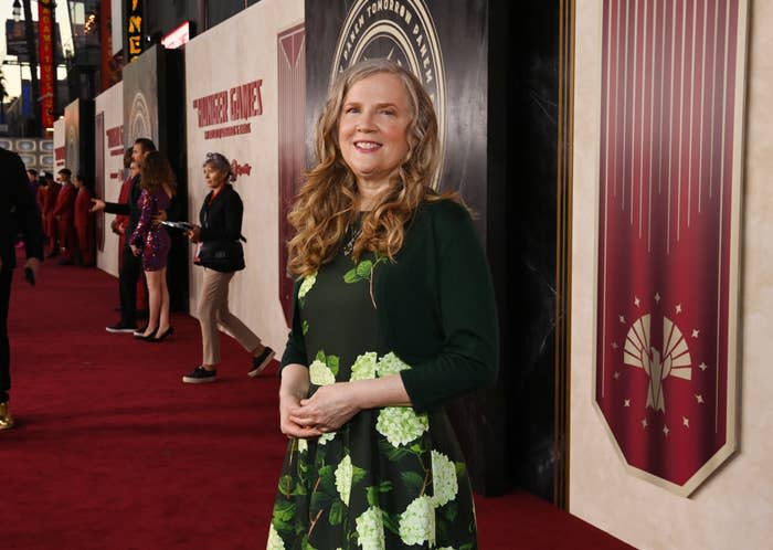 Suzanne Collins stands on the red carpet at a book-related event, wearing a floral dress and a dark jacket. Other attendees and banners are visible in the background