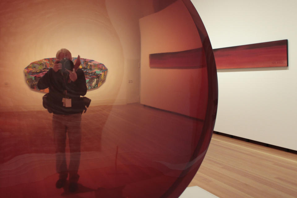 A photographer takes a picture through the sculpture 'Red Concave Circel' from 1970 by artist De Wain Valantine during a press preview of the exhibition 'Pazific Standart Time' in Berlin Wednesday, March 14, 2012. The exhibition that offers viewers a primer on three decades of Los Angeles' diverse art scene and stems from a decade of research is going on show in Berlin from March 15. until June 10, 2012. In the background the painting 'Black Tear' from 1969 by Ronald Davis and right the painting '90% Devil 10% Angel' from 1982 by Ed Ruscha. (AP Photo/Markus Schreiber)