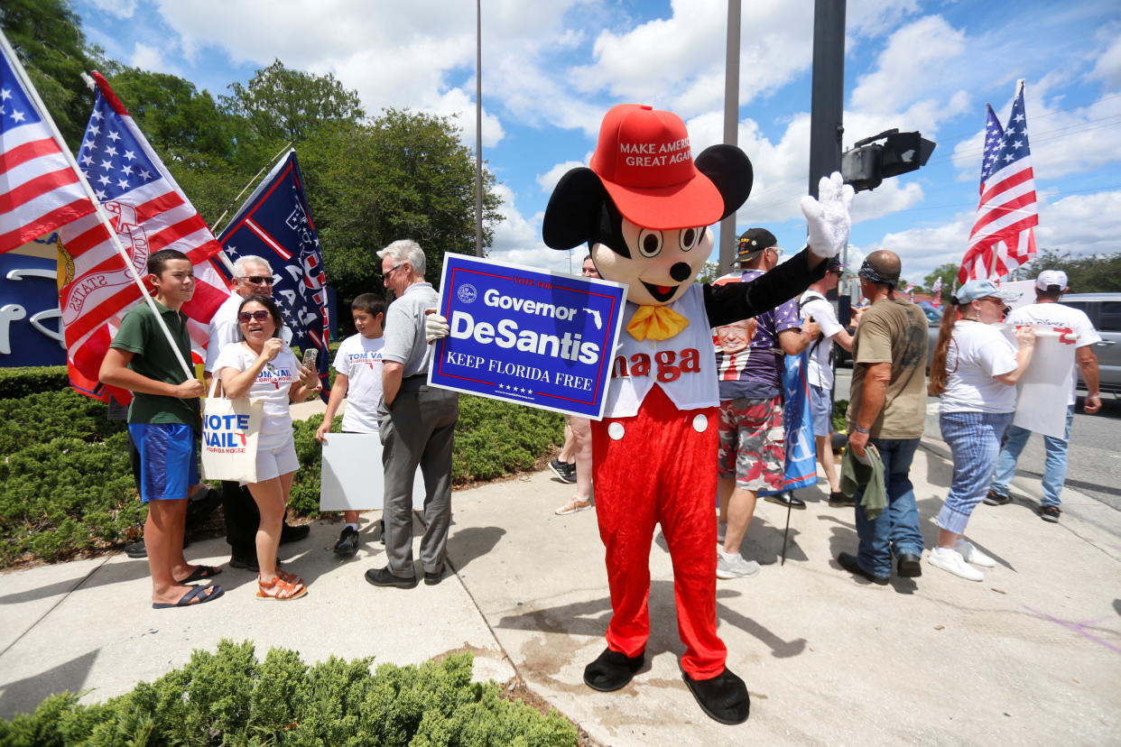 A person wearing a mouse costume holds a Governor Ron DeSantis poster stands where supporters of Florida's Republican-backed 