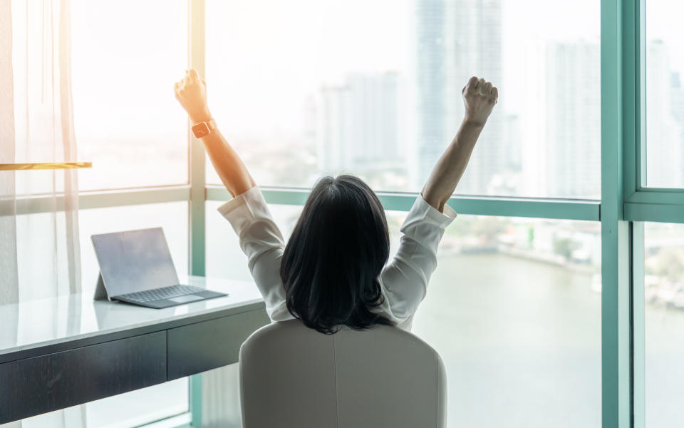 Business achievement concept with happy businesswoman relaxing in office or hotel room, resting and raising fists with ambition looking forward to city building urban scene through glass window