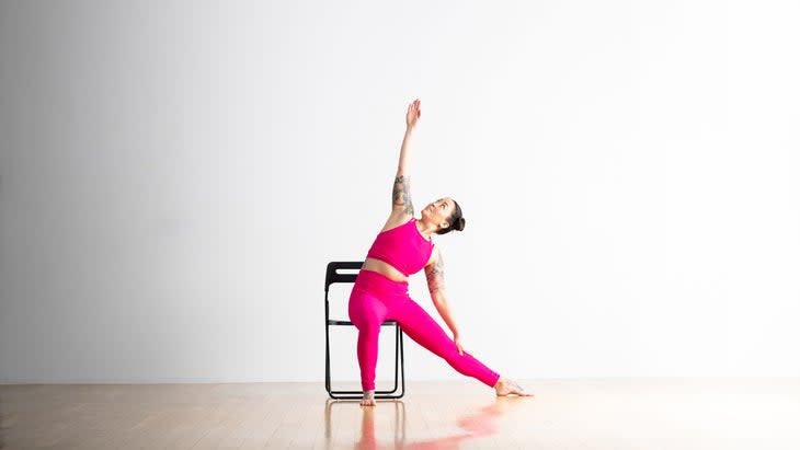 Asian American woman in bright pink top and tights practices Triangle pose in a chair