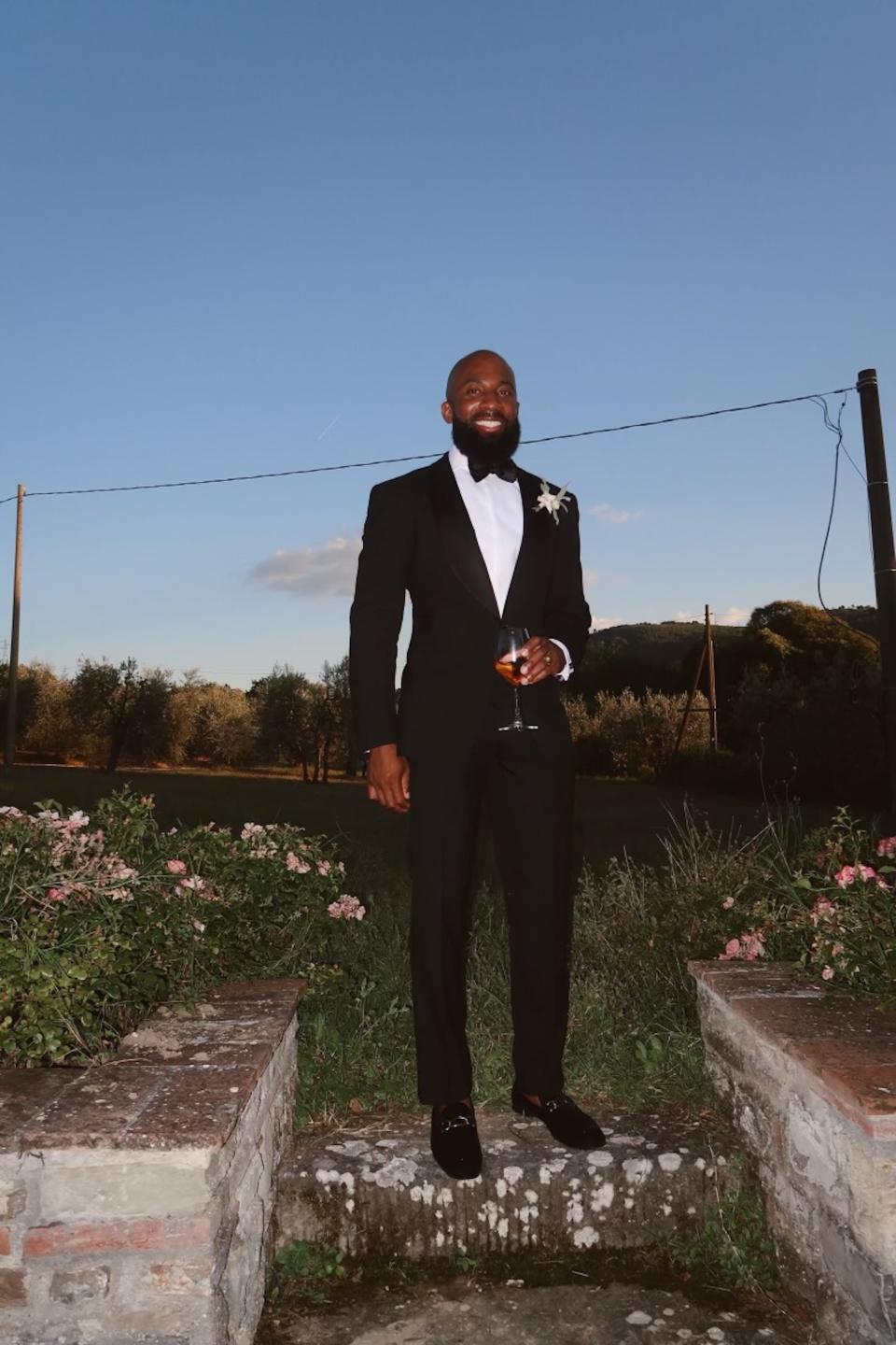 A groom stands in his tuxedo holding a glass of wine.