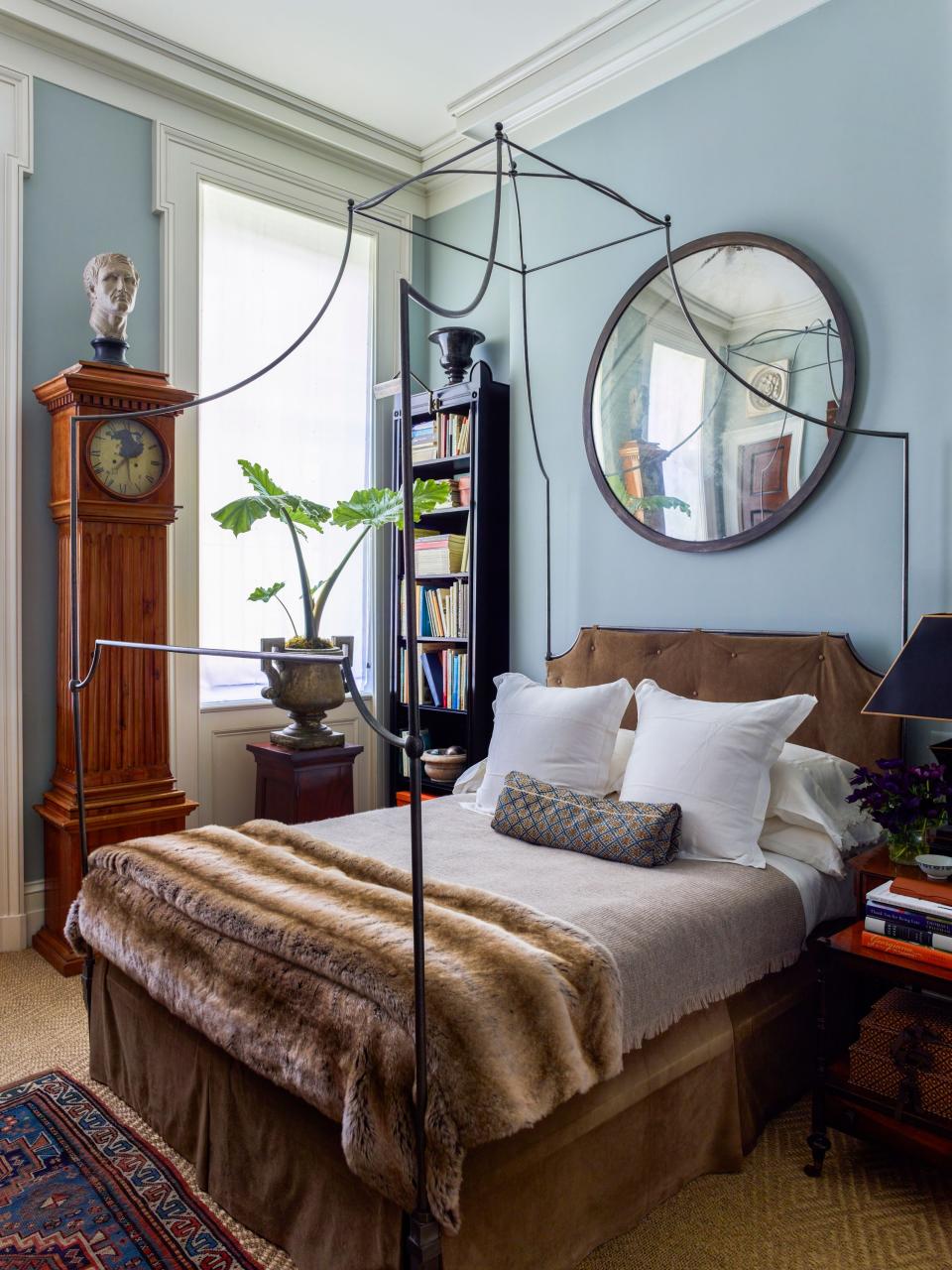 Gil Schafer designed the custom canopy bed in the main suite in the style of a 19th-century campaign bed and had the headboard and bed skirt covered in suede by Dualoy; the faux fur throw is from KRB, and the wool throw is from Aero. He also custom designed the ebonized bookcase in the corner. The antique mirror was purchased at Nadin & Macintosh, the Swedish case clock is from Evergreen Antiques, and the cast-iron urn was bought on a trip to London and placed atop a Regency mahogany pedestal from Cove Landing.