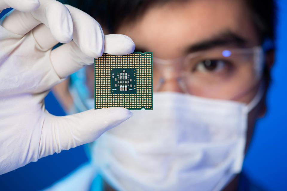 Person wearing face mask, googles, and gloves holding a semiconductor chip in two fingers.