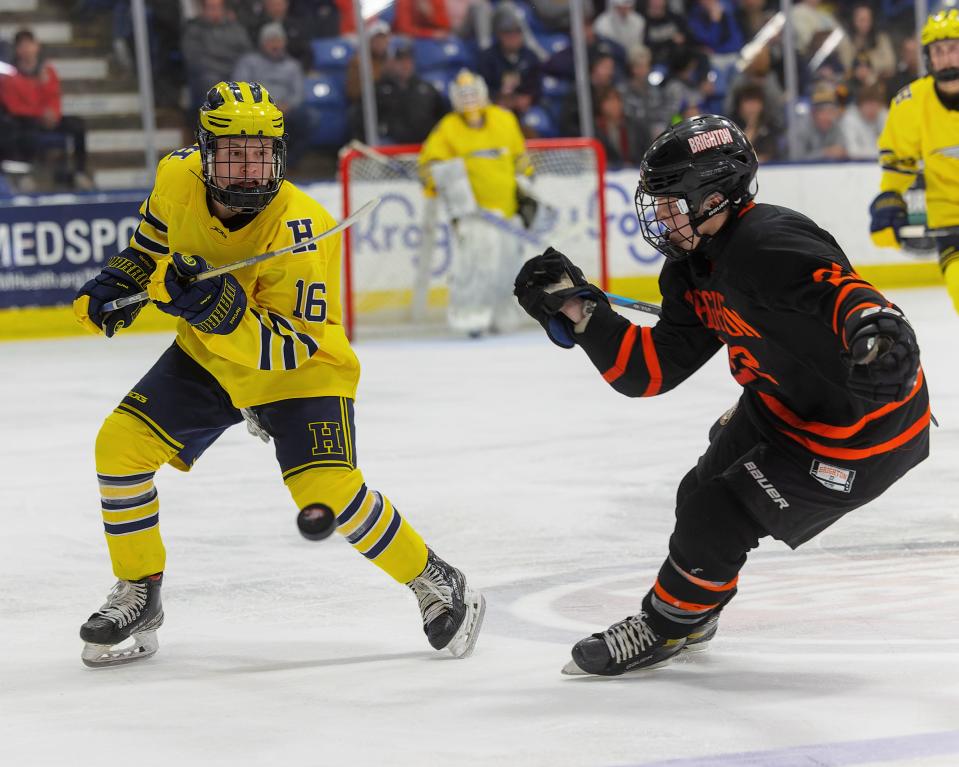 Hartland's Ian Kastamo (16) flips the puck up the ice as Brighton's Lane Petit defends during the Bulldogs' 4-3 triple overtime victory in the state Division 1 semifinals Friday, March, 10, 2023 at USA Hockey Arena.