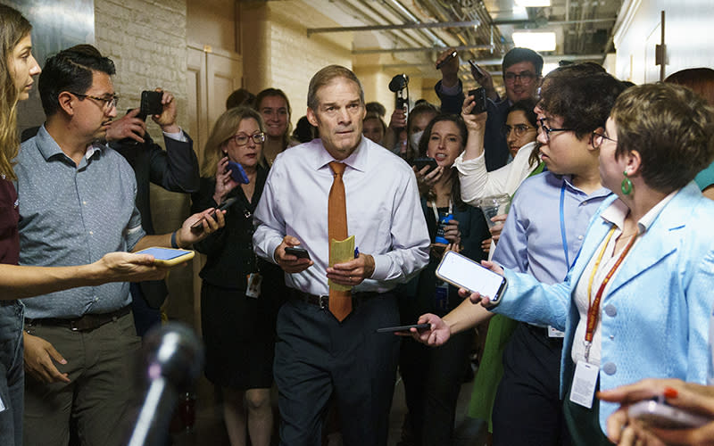 The view is looking down a basement hallway full of people. In the center of the group is Rep. Jim Jordan (R-Ohio). Around him are reporters holding up smartphones and cameras to interview him.