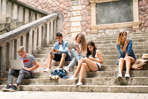 Teenagers with smartphones and computers sit on stone steps.