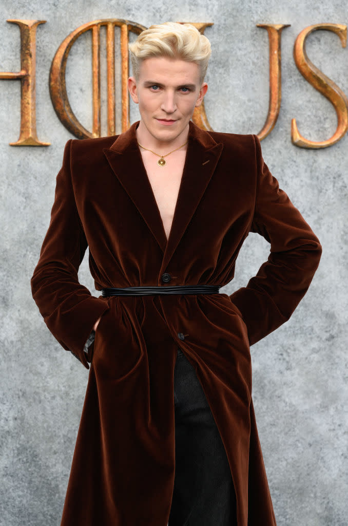 Tom Glynn-Carney poses on the red carpet in a long velvet coat with a belt at the "House of the Dragon" event