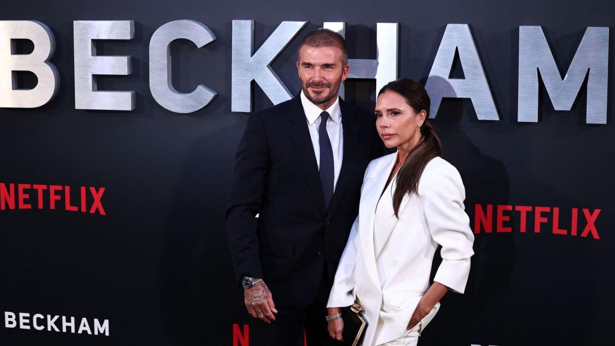 david beckham, wearing a black suit, and victoria beckham, wearing a white dress, standing in front netflix's beckham documentary