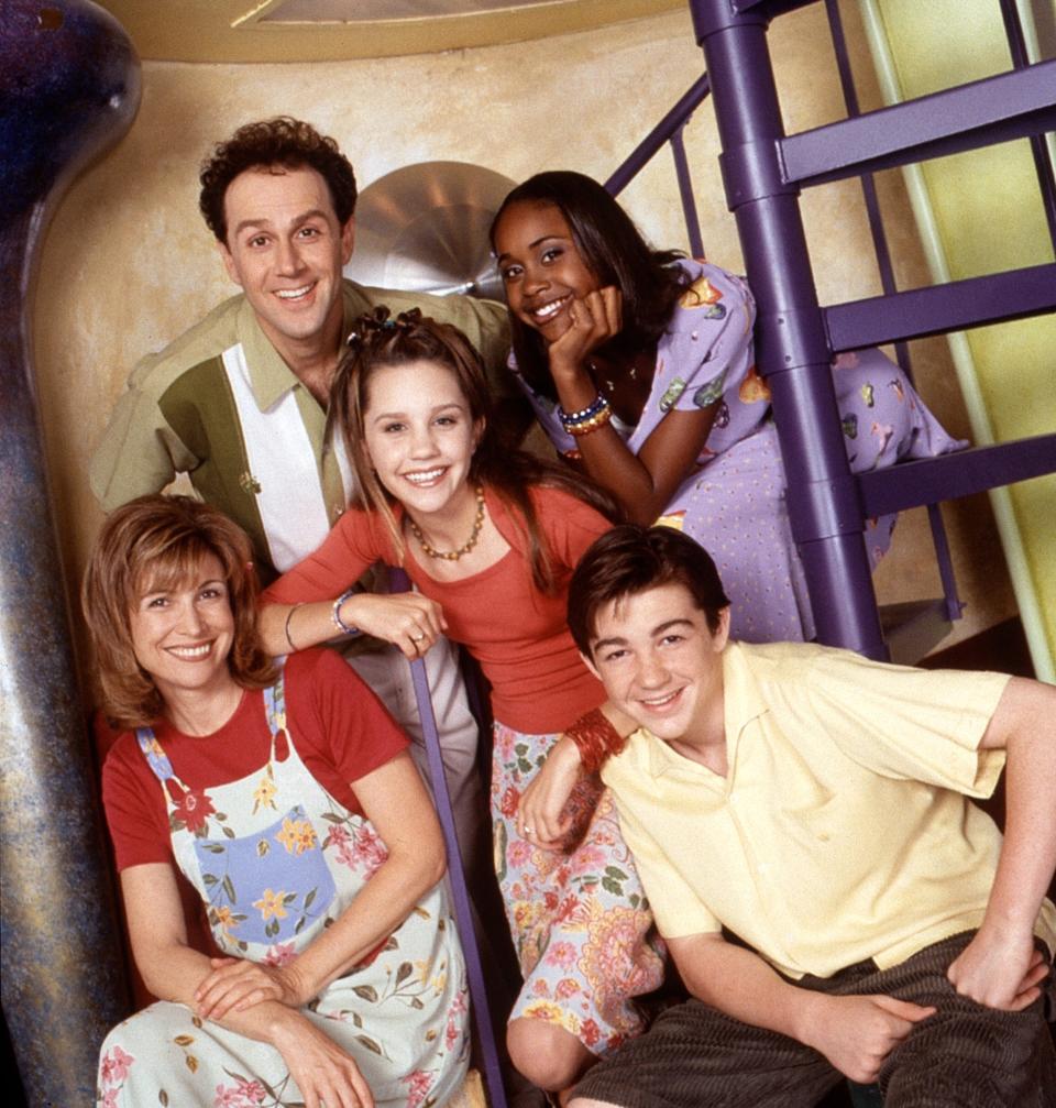 Five cast members from the TV show posing together with a spiral staircase in the background