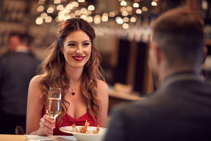 Woman in an elegant red dress with a man, at a table holding a glass, in a restaurant setting