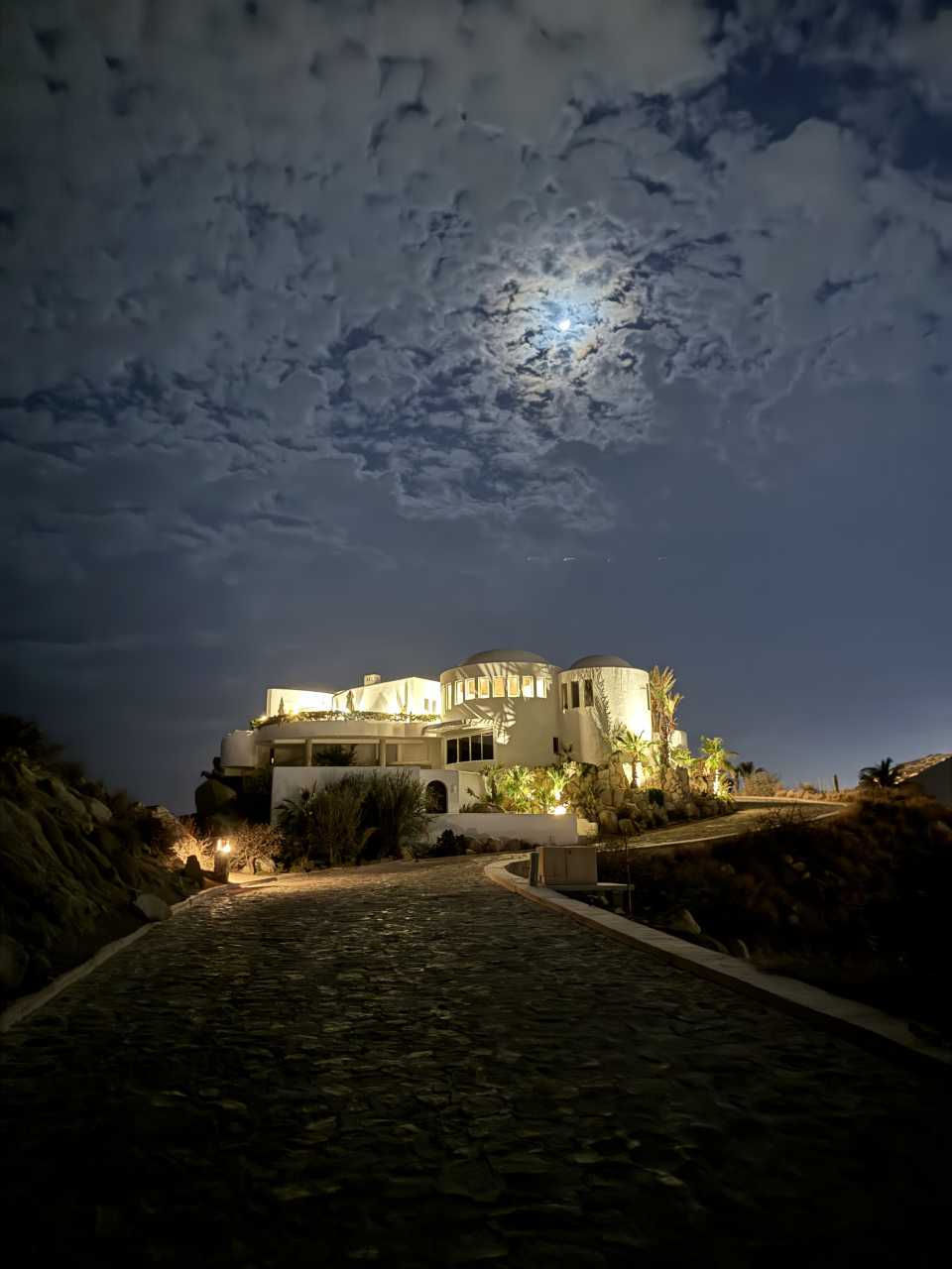 A moonlit path leads to a white building on a hilltop, surrounded by lush vegetation and rocky terrain, under a cloudy night sky. Image fits travel category