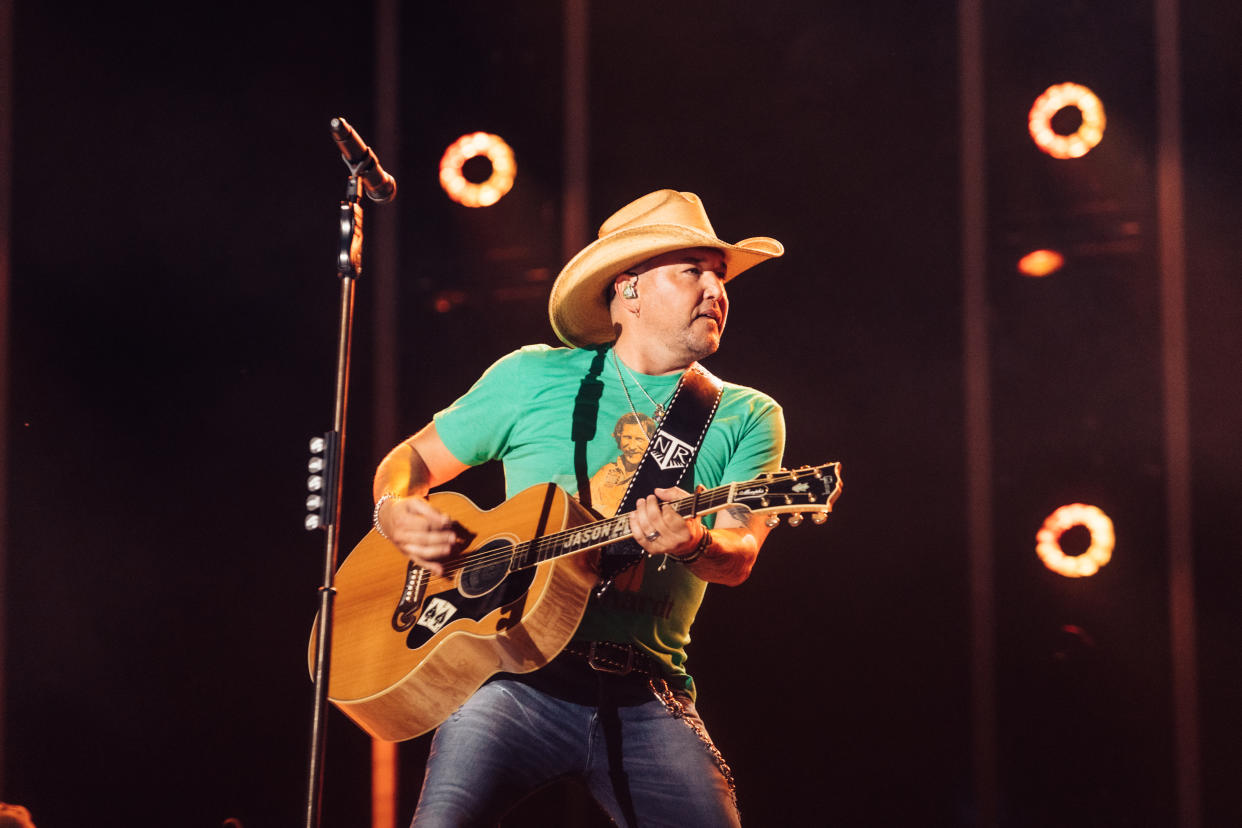 Jason Aldean onstage wearing a cowboy hat and holding an acoustic guitar.