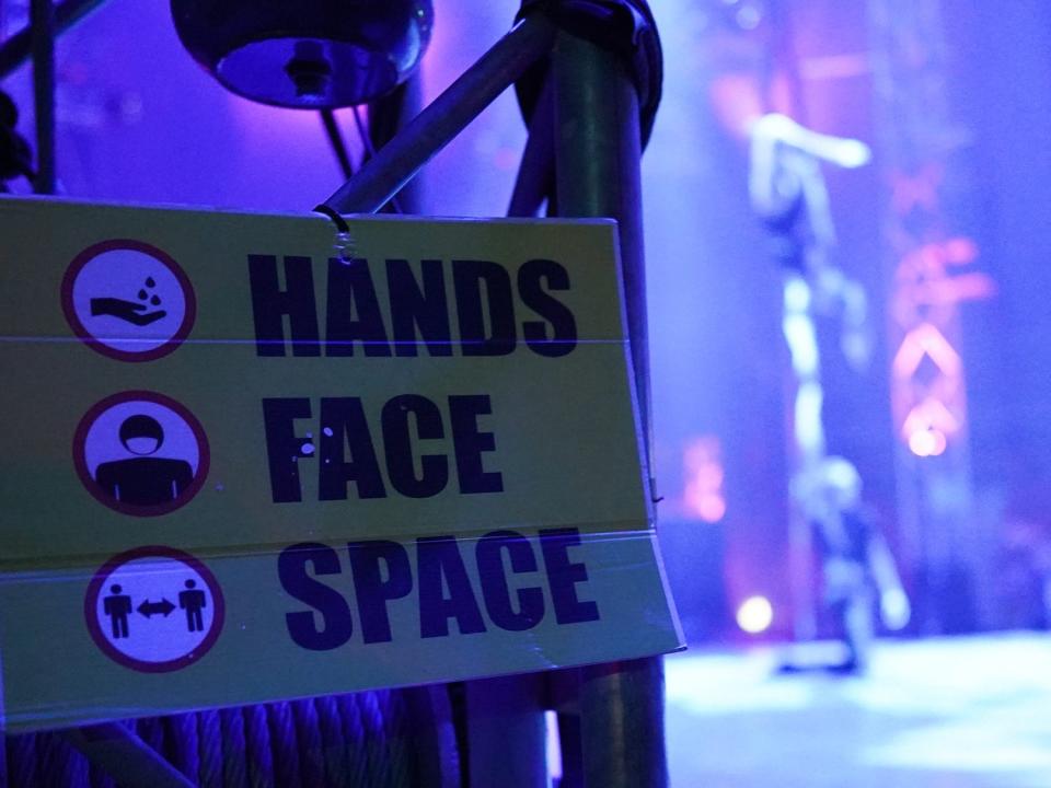 A yellow sign shows the UK's public health messaging "hands, face, space". In the background and circus performers.