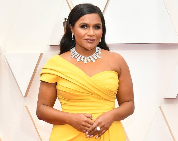 Mindy smiles on a red carpet while wearing a yellow one shoulder dress and a diamond necklace