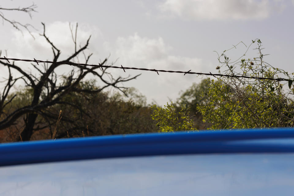 A barbed wire fence line is seen along the side of a roadway where a water station for immigrants containing sealed jugs of fresh water sits in rural Jim Hogg County, Texas, Tuesday, July 25, 2023. The South Texas Human Rights Center maintains over 100 blue barrels consistently stocked with water across rural South Texas to serve as a life-saving measure for immigrants who have crossed into the United States to travel north in the sweltering heat. (AP Photo/Michael Gonzalez)