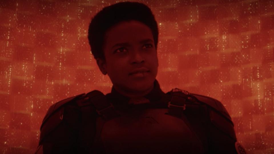 A woman in a black military suit sits in a red room looking stern