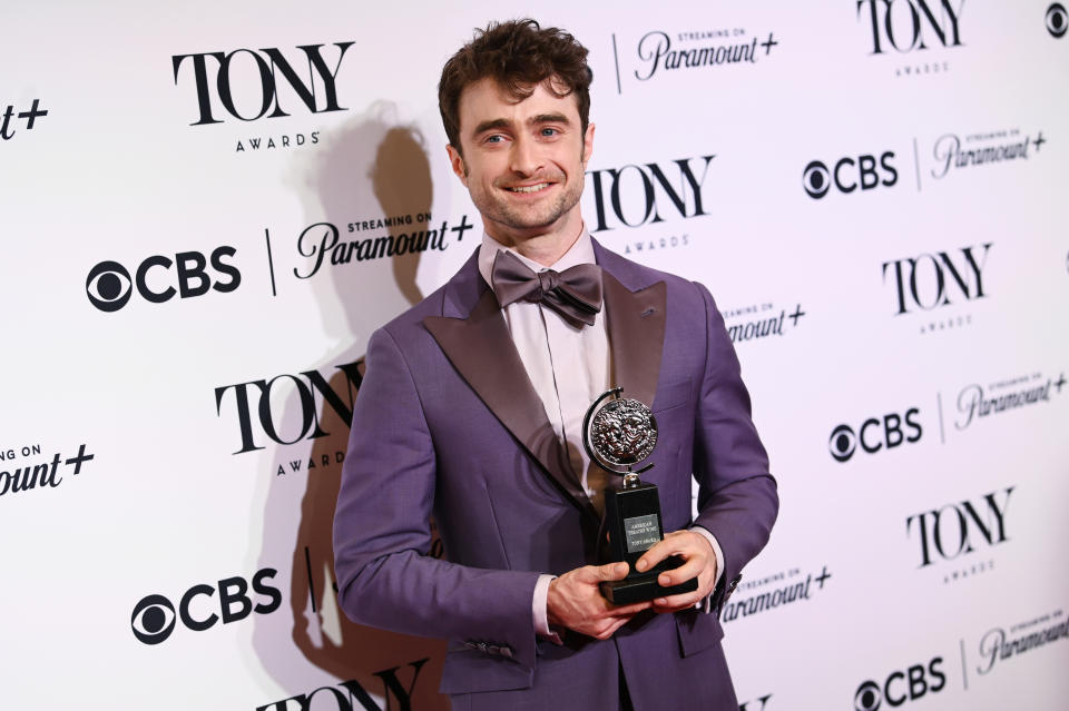 Daniel Radcliffe, in a sharp suit with a bow tie, holds a Tony Award while posing for photos at a Tony Awards event backdrop