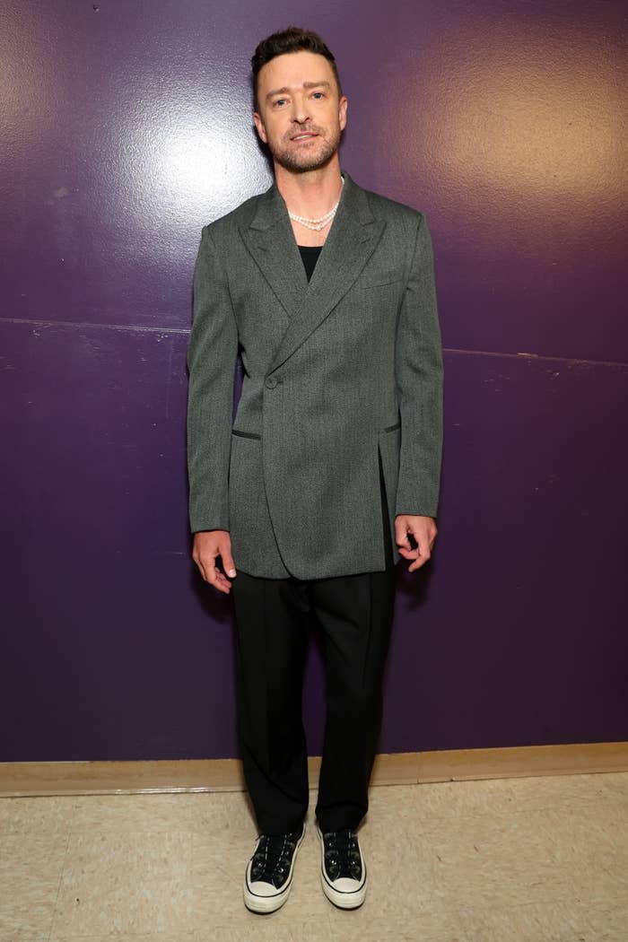 Justin Timberlake stands against a plain wall in a double-breasted blazer, black pants and sneakers.