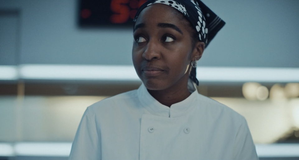 Sydney from The Bear with a patterned headscarf looks off to the side while wearing a double-breasted chef jacket in a professional kitchen setting