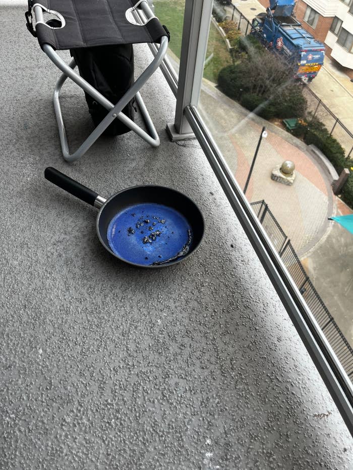 A frying pan with burnt food inside is placed on a balcony floor. The pan's surface has turned blue