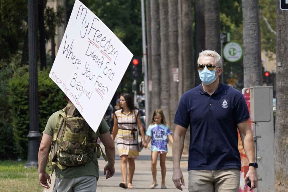 A person toting a sign from a demonstration against mandatory vaccinations passes a man wearing a face mask by the Capitol in Sacramento, Calif., Monday, Aug. 16, 2021. California is mandating COVID-19 vaccinations for all health care workers and will require state government and school staff to be vaccinated or regularly tested.(AP Photo/Rich Pedroncelli)