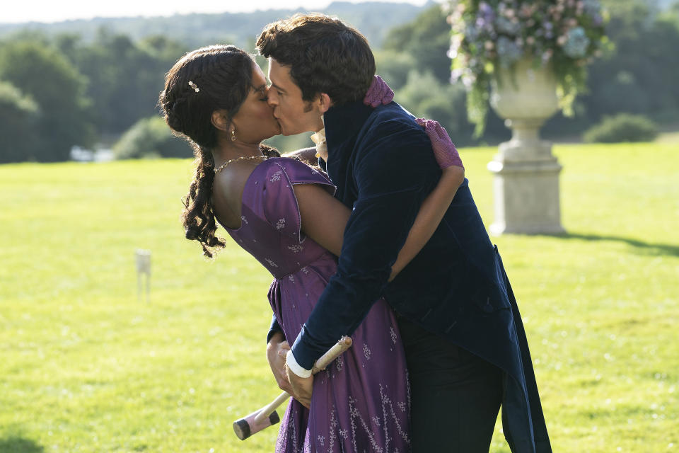 A man in Regency-era coattails and a woman in a purple dress of the same era kiss on a lawn, his arms wrapped around her lower torso and one hand holding a croquet mallet; still from "Bridgerton" Season 2.