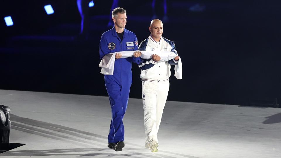  A man in a blue flight suit and a man in a white athletic tracksuit walk on a lighted stage carrying a folded flag. 